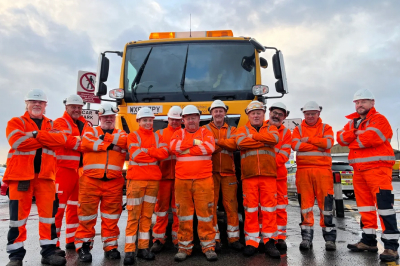 A group of smiling men in high vis clothing stand in front of a gritting truck